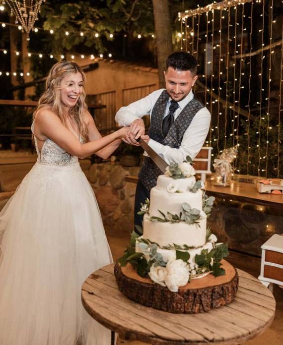 Pareja sonriente cortando juntos una tarta de tres pisos decorada con hojas verdes sobre una mesa de madera rústica.