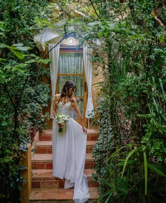 Mujer con vestido blanco y ramo de flores posando en unas escaleras de piedra rodeadas de hiedra y vegetación natural.