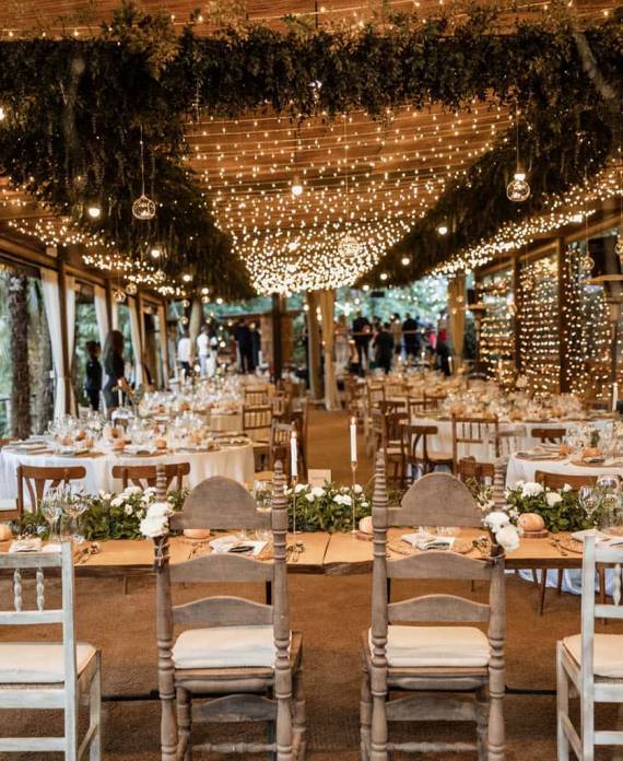 Mesas largas de madera montadas para un banquete bajo una estructura decorada con miles de luces brillantes.