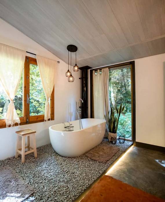 Cuarto de baño moderno con una bañera ovalada blanca sobre suelo de piedra, junto a un ventanal abierto hacia el bosque.