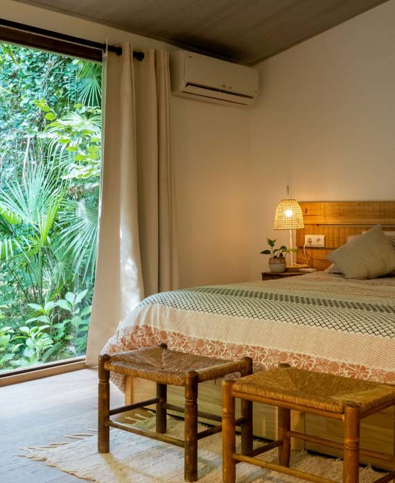 ormitorio con cama doble, mesitas de noche de mimbre y un gran ventanal que da directamente a la vegetación exterior.
